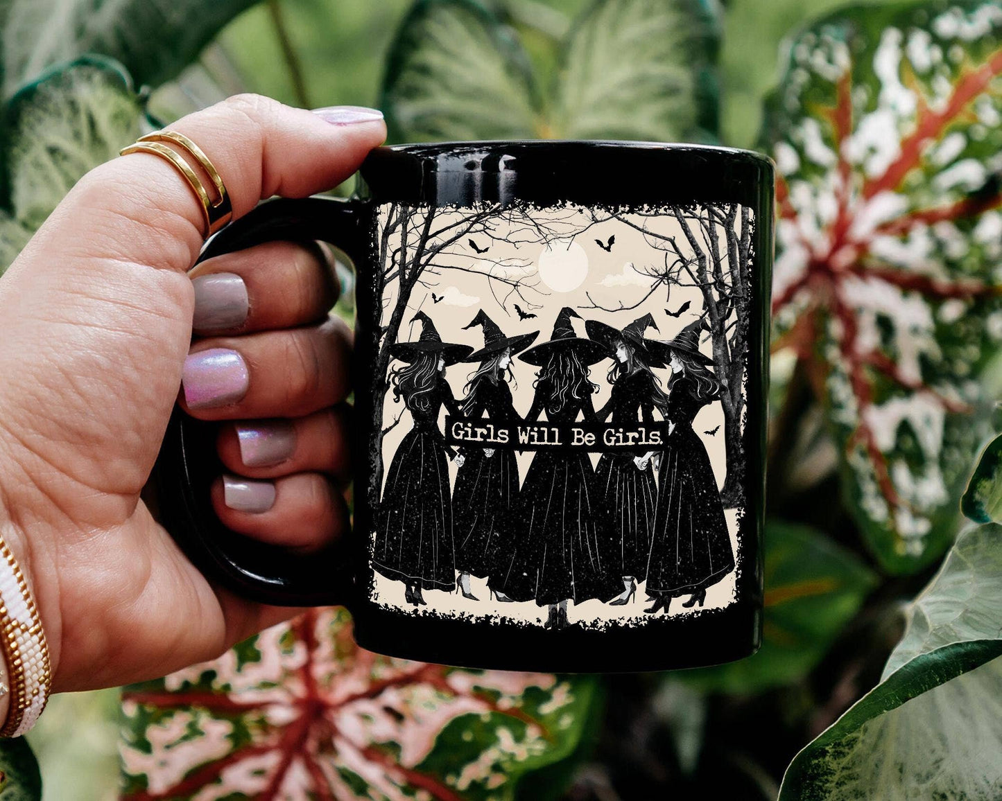 Hand holding a black mug with a design of three women in black dresses and the text 'Girls Will Be Girls' against a natural background.