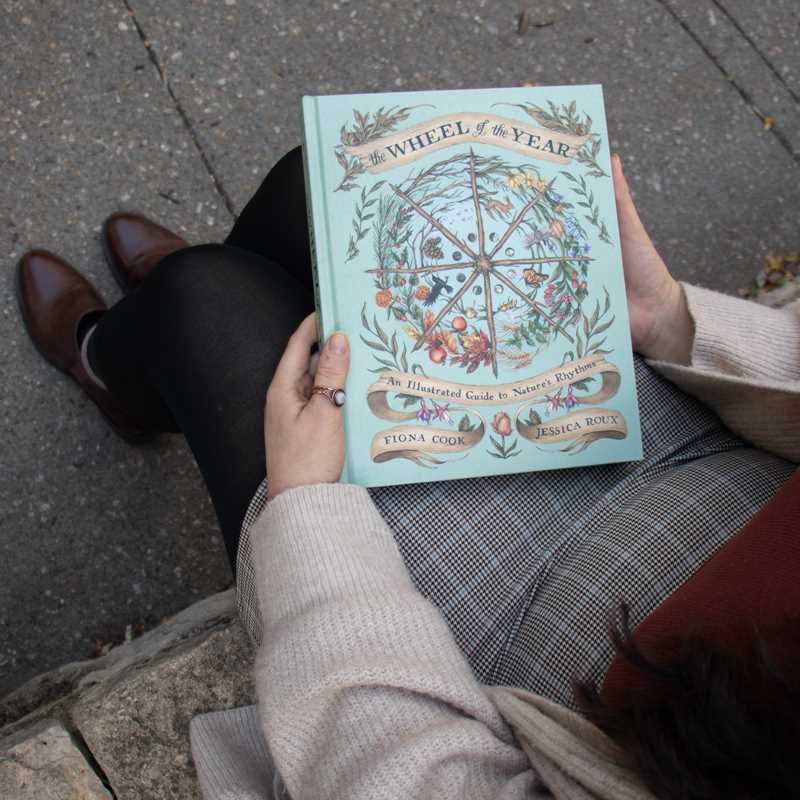 Person holding a book titled 'Wheel of the Year' on a sidewalk.