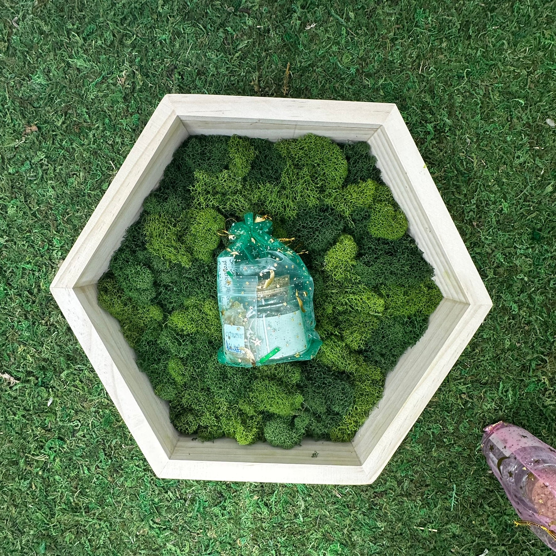 A spell jar kit containing candle and herbs on a bed of green moss, presented on a grass-like surface.