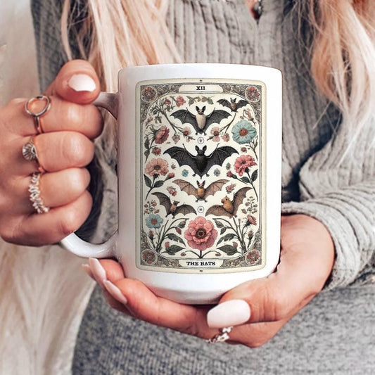 Person holding a mug with a tarot card design featuring bats and flowers.