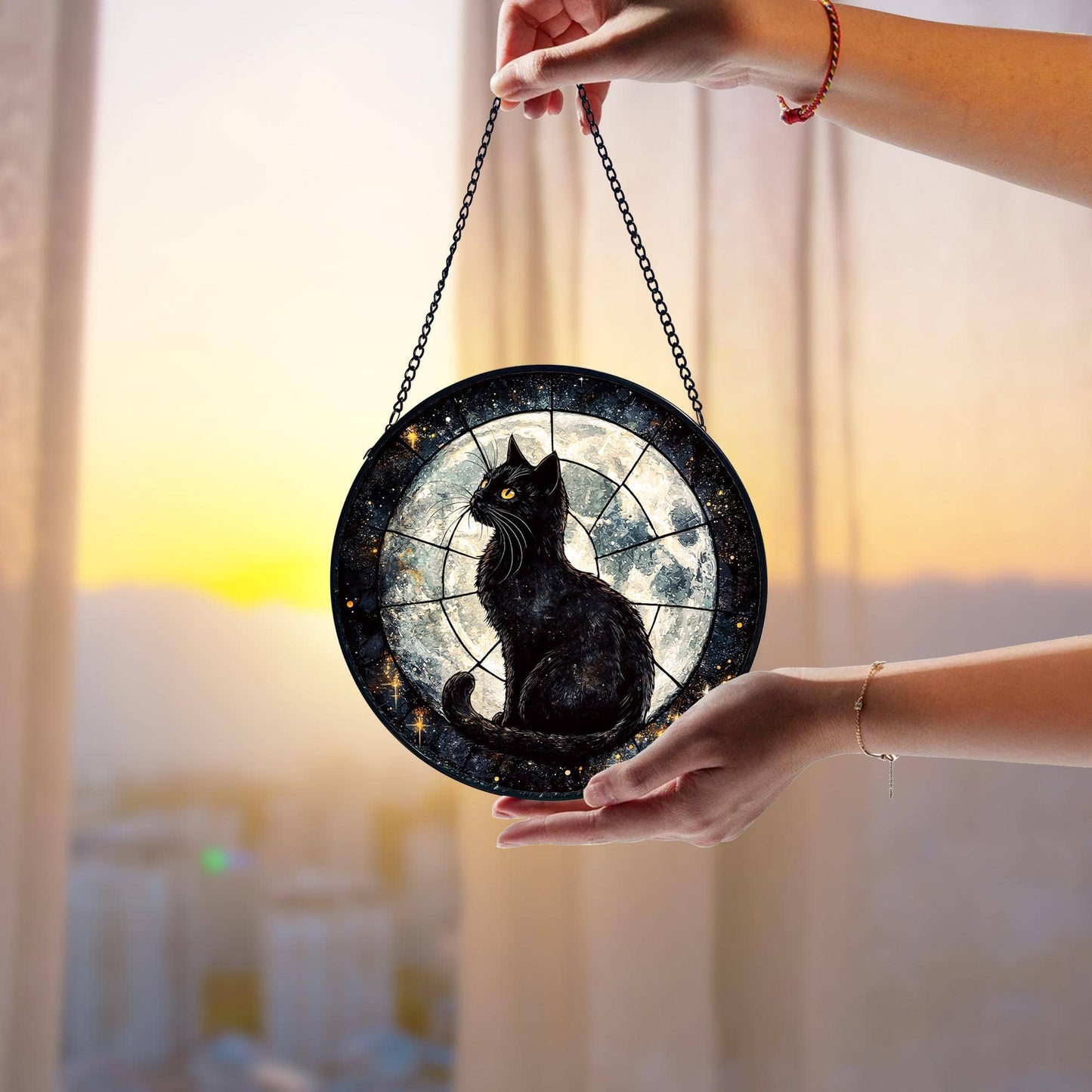 Round decorative item with a black cat design held by a person against a blurred sunset background.
