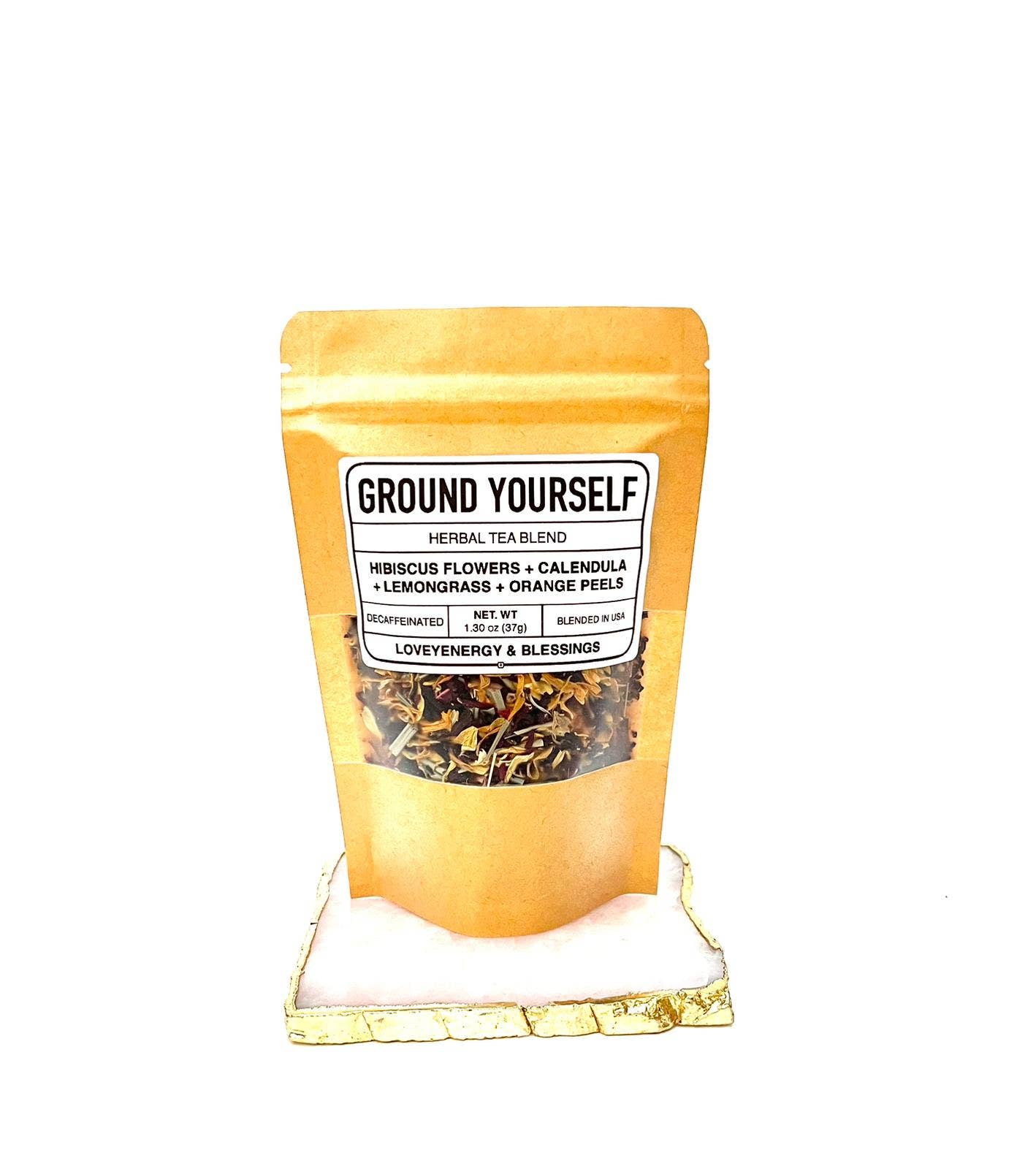 A sealed, gold-colored bag of Ground Yourself handcrafted herbal tea blend, with a label showing the product name and lists of ingredients such as hibiscus flowers, lemongrass, calendula petals, and orange peels.
