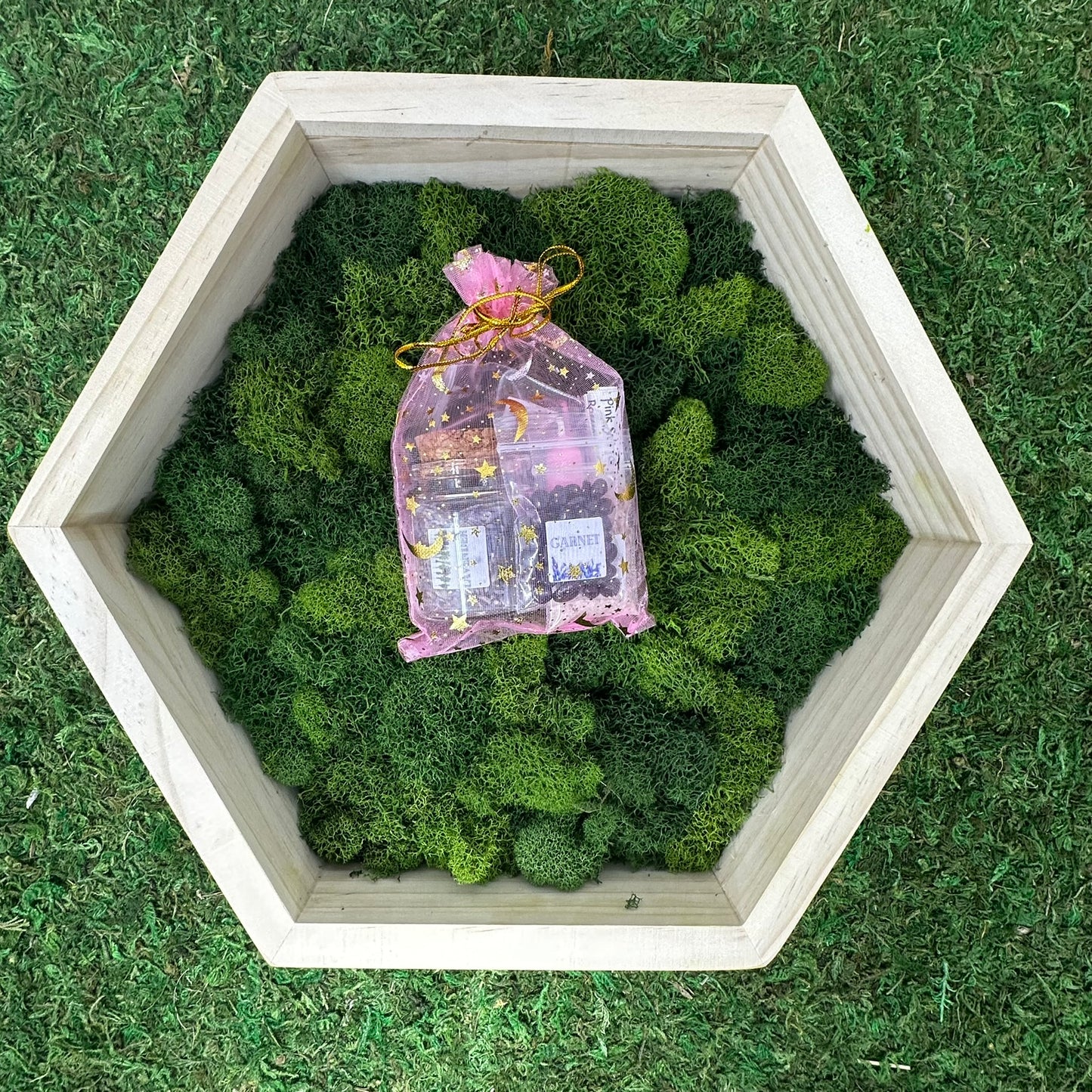 A spell jar kit containing pink candle and herbs on a bed of green moss, presented on a grass-like surface.