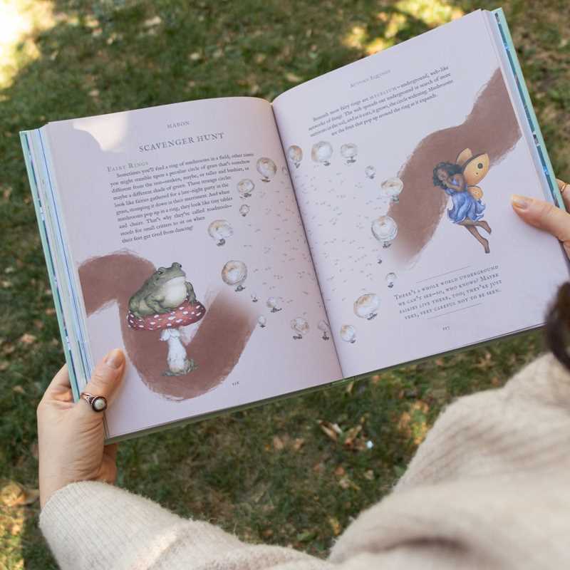 Person holding a children's book with illustrations of a frog and a fairy on a grassy background