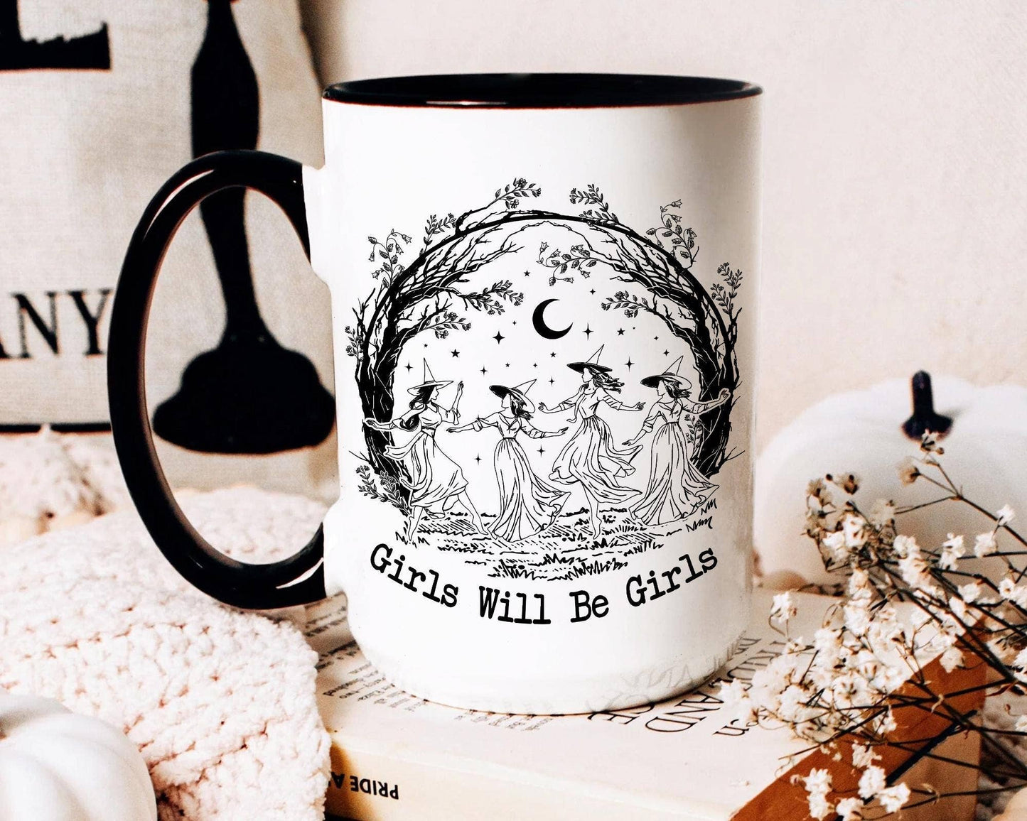 Mug with 'Girls Will Be Girls' design featuring three women and a moon, placed on a surface with decorative items.