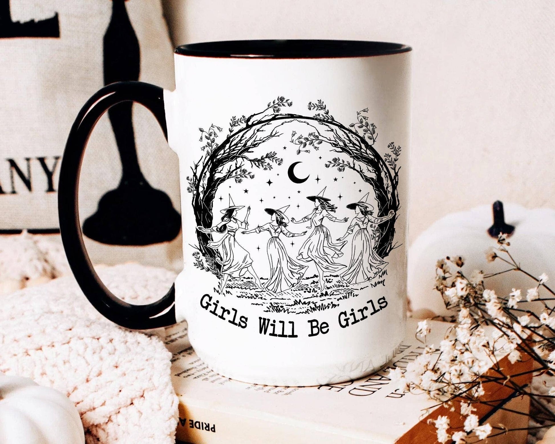 Mug with 'Girls Will Be Girls' design featuring three women and a moon, placed on a surface with decorative items.