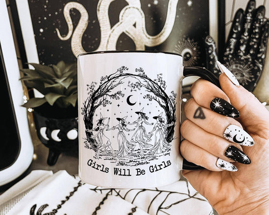 Mug with 'Girls Will Be Girls' design held by a hand with decorative nails, against a mystical background.